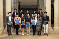 Foto: Eine Gruppe von Personen in akademischen Roben hält Urkunden der Universität Hohenheim. Sie stehen in einem Gebäude mit Säulen und einer Treppe im Hintergrund.