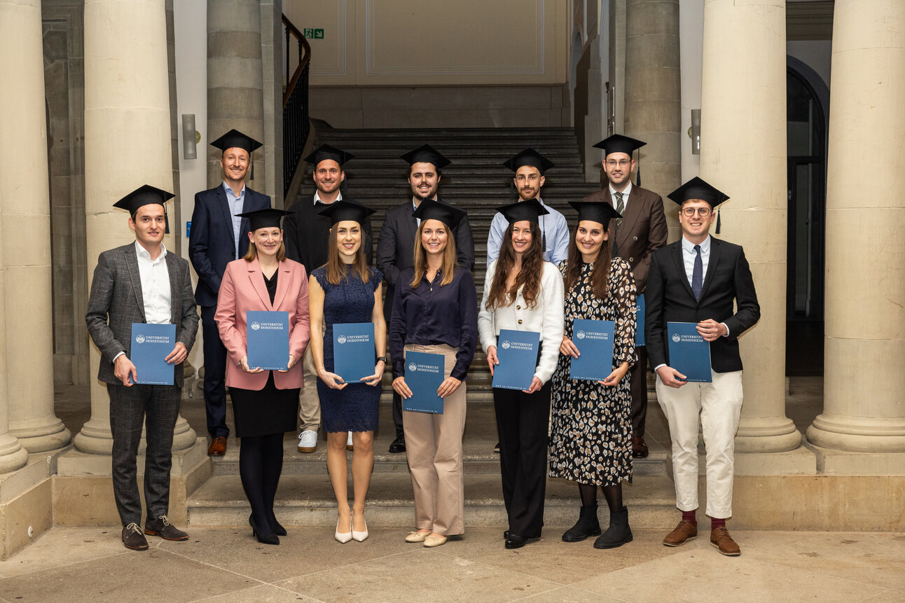 Foto: Eine Gruppe von Personen in akademischen Roben hält Urkunden der Universität Hohenheim. Sie stehen in einem Gebäude mit Säulen und einer Treppe im Hintergrund.