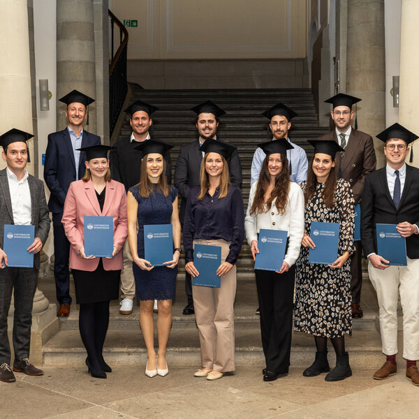 Foto: Eine Gruppe von Personen in akademischen Roben hält Urkunden der Universität Hohenheim. Sie stehen in einem Gebäude mit Säulen und einer Treppe im Hintergrund.