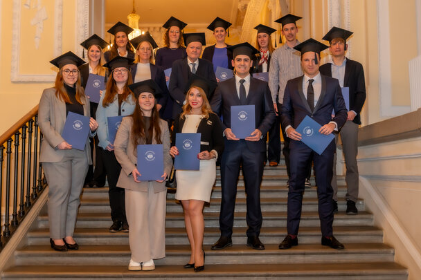 Foto: Eine Gruppe von Personen in formeller Kleidung steht auf einer Treppe. Sie tragen alle schwarze Hüte und halten blaue Urkundenmappen mit dem Logo der Universität Hohenheim.