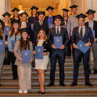 Foto: Eine Gruppe von Personen in formeller Kleidung steht auf einer Treppe. Sie tragen alle schwarze Hüte und halten blaue Urkundenmappen mit dem Logo der Universität Hohenheim.