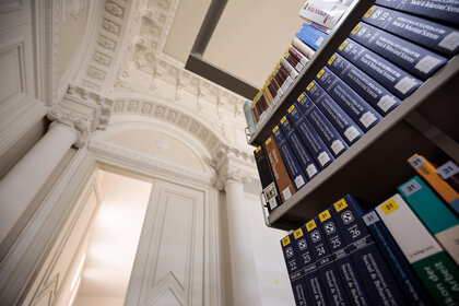 Foto: Ein Bücherregal mit zahlreichen Bänden steht in einer Bibliothek mit aufwendiger klassischer Architektur, einschließlich Säulen und dekorativer Decke.