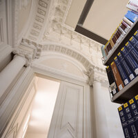 Foto: Ein Bücherregal mit zahlreichen Bänden steht in einer Bibliothek mit aufwendiger klassischer Architektur, einschließlich Säulen und dekorativer Decke.