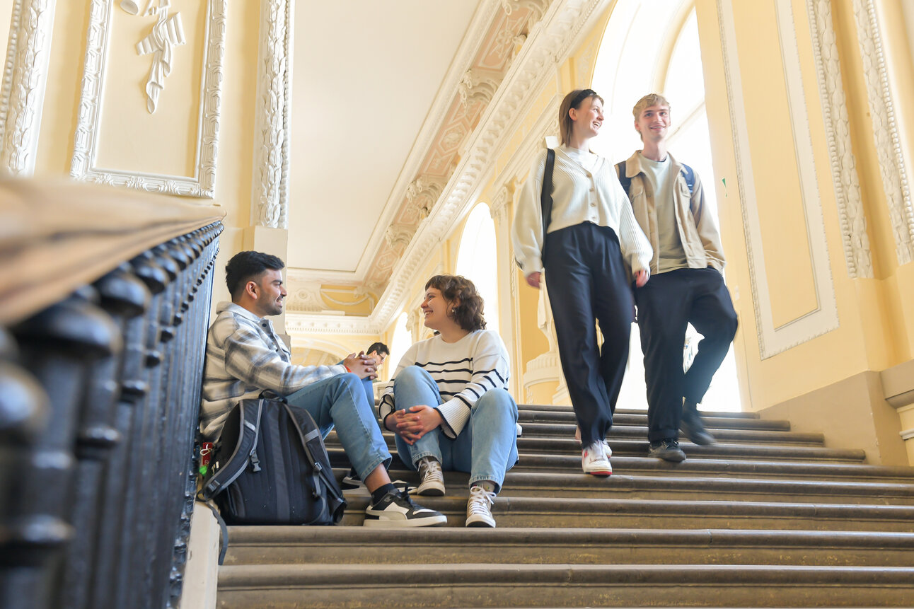 Foto: Mehrere Personen sitzen und stehen auf einer breiten Treppe in einem Gebäude mit dekorativen Wänden und hohen Decken. Zwei Personen sitzen auf den Stufen, während zwei andere die Treppe hinuntergehen.