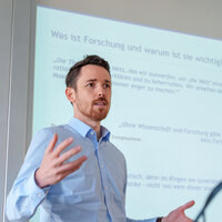 Foto: Eine Person in einem blauen Hemd hält eine Präsentation vor einer Projektionsfläche. Der Text auf der Folie lautet: 'Was ist Forschung und warum ist sie wichtig?' mit Zitaten von Sir Karl Popper. Das Logo der Universität Hohenheim ist sichtbar.