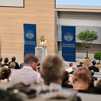 Foto: Eine Person steht an einem Rednerpult im Hörsaal der Universität Hohenheim. Im Hintergrund sind zwei Banner mit dem Logo der Universität sichtbar. Das Publikum sitzt und hört zu.