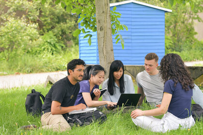 Foto: Eine Gruppe von Personen sitzt auf einer Wiese unter einem Baum. Sie sind um einen Laptop versammelt und scheinen zu arbeiten oder zu lernen. Im Hintergrund ist ein blaues Gebäude sichtbar.