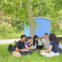 Foto: Eine Gruppe von Personen sitzt auf einer Wiese unter einem Baum. Sie sind um einen Laptop versammelt und scheinen zu arbeiten oder zu lernen. Im Hintergrund ist ein blaues Gebäude sichtbar.