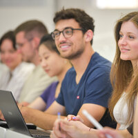 Foto: Eine Gruppe von Personen sitzt in einem Hörsaal. Einige haben Laptops und Stifte vor sich. Der Fokus liegt auf einer Person mit einem blauen T-Shirt, die aufmerksam nach vorne schaut.