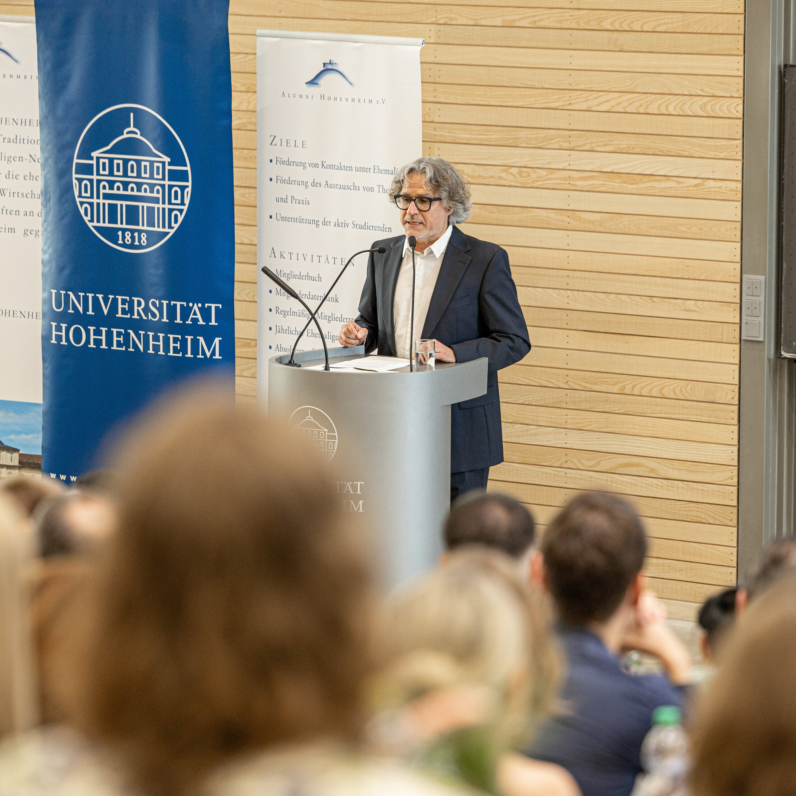 Foto: Eine Person hält einen Vortrag an einem Rednerpult mit dem Logo der Universität Hohenheim. Im Hintergrund sind Banner mit Informationen über Alumni und Ziele der Universität zu sehen. Ein Publikum hört zu.