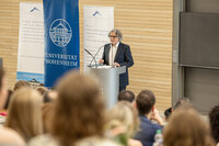 Foto: Eine Person hält einen Vortrag an einem Rednerpult mit dem Logo der Universität Hohenheim. Im Hintergrund sind Banner mit Informationen über Alumni und Ziele der Universität zu sehen. Ein Publikum hört zu.