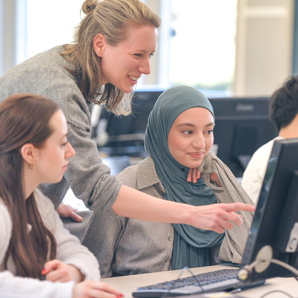 Foto: Mehrere Personen sitzen an Computern in einem Kursraum. Eine Person zeigt auf den Bildschirm eines Computers, während andere aufmerksam zuschauen. Die Umgebung wirkt modern und professionell.