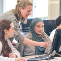 Foto: Mehrere Personen sitzen an Computern in einem Kursraum. Eine Person zeigt auf den Bildschirm eines Computers, während andere aufmerksam zuschauen. Die Umgebung wirkt modern und professionell.