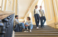 Foto: Mehrere Personen sitzen und stehen auf einer breiten Treppe in einem Gebäude mit dekorativen Wänden und hohen Decken. Zwei Personen sitzen auf den Stufen, während zwei andere die Treppe hinuntergehen.