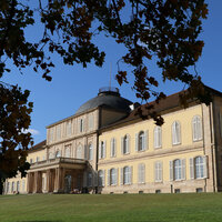Foto: Schloss Hohenheim mit symmetrischem Design und Kuppeldach, umgeben von herbstlichen Bäumen und einem gepflegten Rasen unter blauem Himmel.