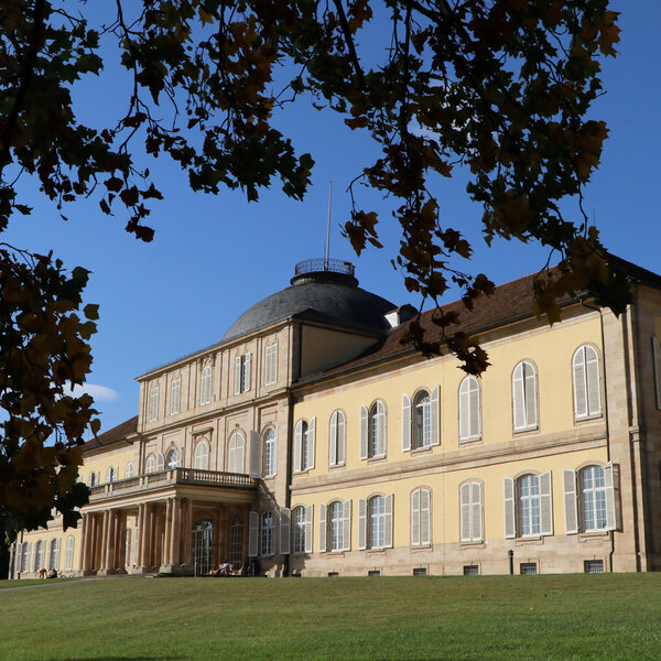 Foto: Schloss Hohenheim mit symmetrischem Design und Kuppeldach, umgeben von herbstlichen Bäumen und einem gepflegten Rasen unter blauem Himmel.