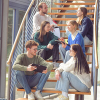 Foto: Eine Gruppe von Personen sitzt auf einer Holztreppe in einem modernen Gebäude. Sie halten Tablets und Notizblöcke und scheinen in einer Diskussion vertieft zu sein.