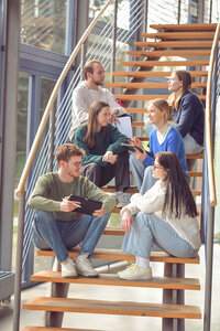 Foto: Eine Gruppe von Personen sitzt auf einer Holztreppe in einem modernen Gebäude. Sie halten Tablets und Notizblöcke und scheinen in einer Diskussion vertieft zu sein.