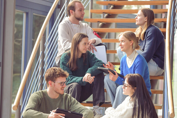 Foto: Eine Gruppe von Personen sitzt auf einer Holztreppe in einem modernen Gebäude. Sie halten Tablets und Notizblöcke und scheinen in einer Diskussion vertieft zu sein.