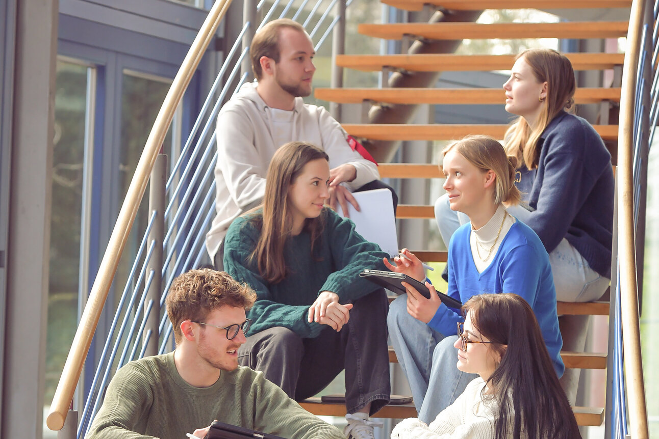 Foto: Eine Gruppe von Personen sitzt auf einer Holztreppe in einem modernen Gebäude. Sie halten Tablets und Notizblöcke und scheinen in einer Diskussion vertieft zu sein.
