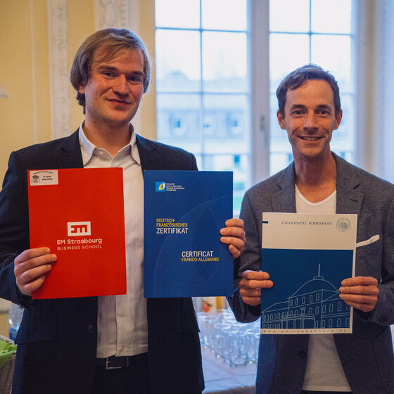 Foto: Zwei Personen halten Zertifikate. Links ein rotes Zertifikat der EM Strasbourg Business School, in der Mitte ein blaues deutsch-französisches Zertifikat, rechts ein Zertifikat der Universität Hohenheim.