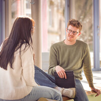 Foto: Zwei Personen sitzen auf einer Fensterbank und unterhalten sich. Eine Person hält ein Tablet. Im Hintergrund sind große Fenster zu sehen, durch die Tageslicht einfällt.