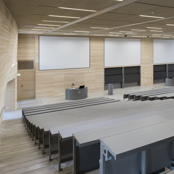Foto: Moderner Hörsaal mit leeren Sitzreihen, großen Projektionsflächen und einem Rednerpult. Die Wände sind aus hellem Holz, und die Decke hat integrierte Beleuchtung.