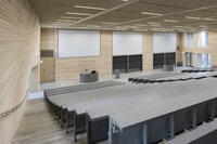 Foto: Moderner Hörsaal mit leeren Sitzreihen, großen Projektionsflächen und einem Rednerpult. Die Wände sind aus hellem Holz, und die Decke hat integrierte Beleuchtung.