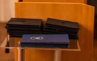 Foto: Stapel von schwarzen Abschlusskappen und blauen Zertifikaten mit dem Logo der Universität Hohenheim auf einem Tisch.