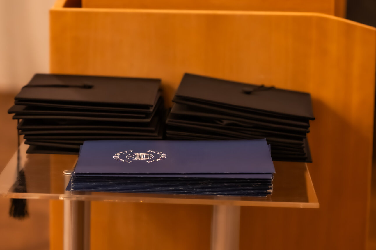 Foto: Stapel von schwarzen Abschlusskappen und blauen Zertifikaten mit dem Logo der Universität Hohenheim auf einem Tisch.