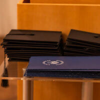 Foto: Stapel von schwarzen Abschlusskappen und blauen Zertifikaten mit dem Logo der Universität Hohenheim auf einem Tisch.