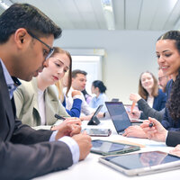 Foto: Eine Gruppe von Personen sitzt an einem Tisch in einem Büro und verwendet Tablets und Laptops. Sie scheinen in ein Gespräch vertieft zu sein, während sie Notizen machen und auf die Geräte schauen.