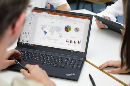 Foto: Eine Person arbeitet an einem Laptop mit einer Präsentation, die Diagramme und eine Weltkarte zeigt. Zwei weitere Personen sind im Hintergrund mit Tablets sichtbar.