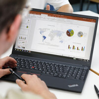 Foto: Eine Person arbeitet an einem Laptop mit einer Präsentation, die Diagramme und eine Weltkarte zeigt. Zwei weitere Personen sind im Hintergrund mit Tablets sichtbar.