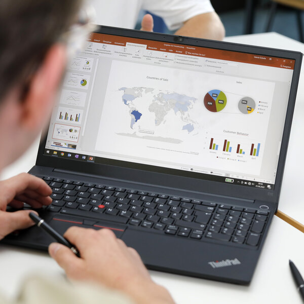Foto: Eine Person arbeitet an einem Laptop mit einer Präsentation, die Diagramme und eine Weltkarte zeigt. Zwei weitere Personen sind im Hintergrund mit Tablets sichtbar.