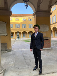 Foto: Eine Person in einem dunklen Anzug steht in einem historischen Innenhof mit gelben Gebäuden und Arkaden. Im Hintergrund sind Fenster mit blauen Fensterläden und eine Statue sichtbar.