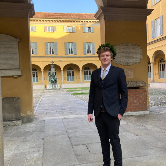 Foto: Eine Person in einem dunklen Anzug steht in einem historischen Innenhof mit gelben Gebäuden und Arkaden. Im Hintergrund sind Fenster mit blauen Fensterläden und eine Statue sichtbar.