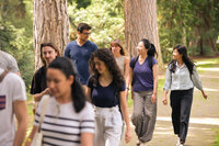 Foto: Eine Gruppe von Personen geht auf einem gepflasterten Weg durch einen bewaldeten Park. Sie tragen Freizeitkleidung und einige haben Rucksäcke. Die Umgebung ist grün und von Bäumen gesäumt.