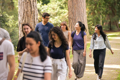 Foto: Eine Gruppe von Personen geht auf einem gepflasterten Weg durch einen bewaldeten Park. Sie tragen Freizeitkleidung und einige haben Rucksäcke. Die Umgebung ist grün und von Bäumen gesäumt.