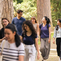 Foto: Eine Gruppe von Personen geht auf einem gepflasterten Weg durch einen bewaldeten Park. Sie tragen Freizeitkleidung und einige haben Rucksäcke. Die Umgebung ist grün und von Bäumen gesäumt.