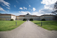Foto: Schloss Hohenheim mit symmetrischem Design und Kuppeldach, umgeben von gepflegtem Rasen unter blauem Himmel mit Wolken. Zwei Personen gehen auf dem Weg zum Eingang.