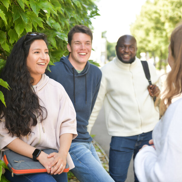 Foto: Eine Gruppe von Personen steht im Freien vor einer Hecke und unterhält sich. Eine Person hält eine Tasche, während eine andere einen Rucksack trägt. Die Umgebung ist grün und sonnig.