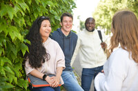 Foto: Eine Gruppe von Personen steht im Freien vor einer Hecke und unterhält sich. Eine Person hält eine Tasche, während eine andere einen Rucksack trägt. Die Umgebung ist grün und sonnig.