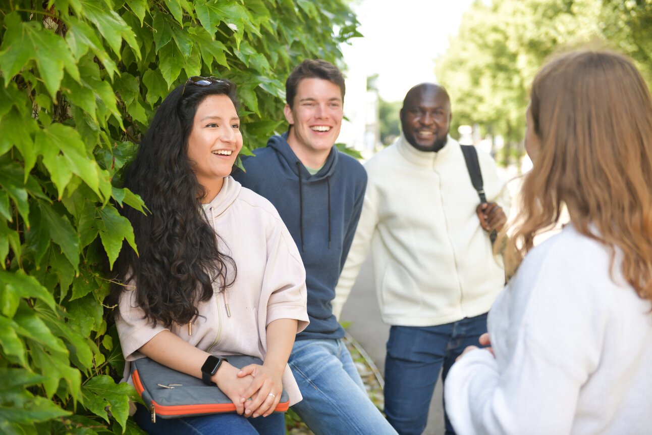 Foto: Eine Gruppe von Personen steht im Freien vor einer Hecke und unterhält sich. Eine Person hält eine Tasche, während eine andere einen Rucksack trägt. Die Umgebung ist grün und sonnig.