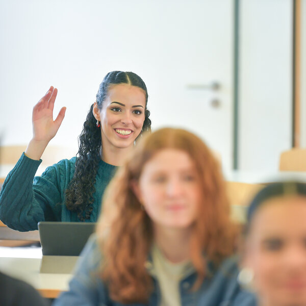 Foto: Mehrere Personen sitzen in einem Klassenzimmer. Eine Person hebt die Hand, während andere aufmerksam sind. Im Hintergrund sind Tische und Stühle sichtbar.