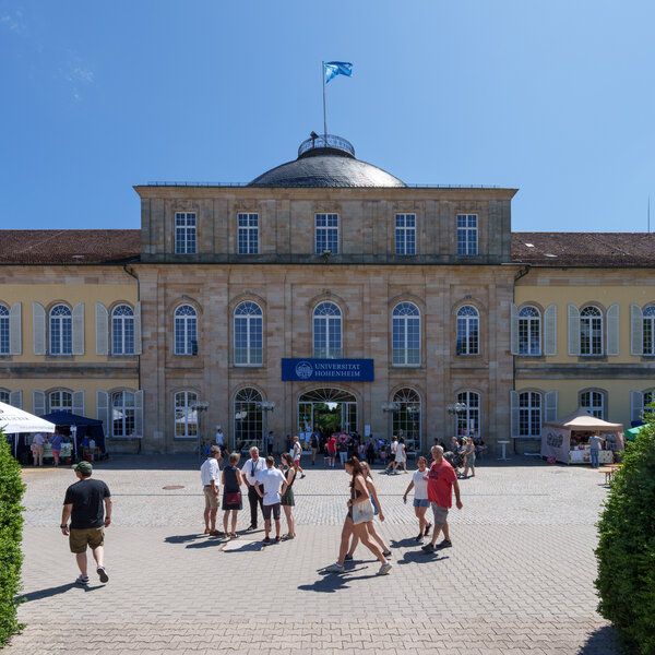 Foto: Schloss Hohenheim mit einer Gruppe von Personen im Vordergrund. Das Gebäude zeigt klassische Architektur mit symmetrischem Design und Kuppeldach. Ein Banner mit der Aufschrift 'Universität Hohenheim' ist sichtbar.