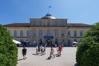 Foto: Schloss Hohenheim mit einer Gruppe von Personen im Vordergrund. Das Gebäude zeigt klassische Architektur mit symmetrischem Design und Kuppeldach. Ein Banner mit der Aufschrift 'Universität Hohenheim' ist sichtbar.