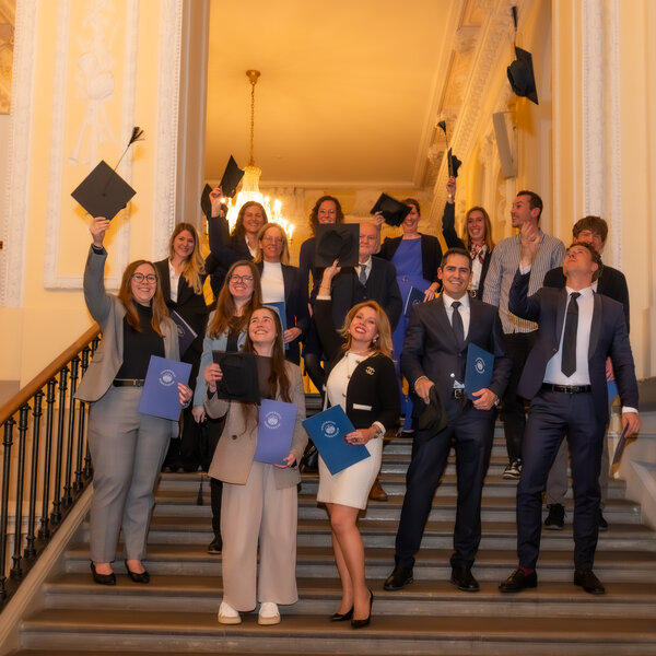 Foto: Eine Gruppe von Personen in formeller Kleidung steht auf einer Treppe in einem Gebäude mit dekorativen Wänden. Einige halten blaue Mappen und werfen Hüte in die Luft.