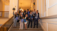 Foto: Eine Gruppe von Personen in formeller Kleidung steht auf einer Treppe in einem Gebäude mit dekorativen Wänden. Einige halten blaue Mappen und werfen Hüte in die Luft.