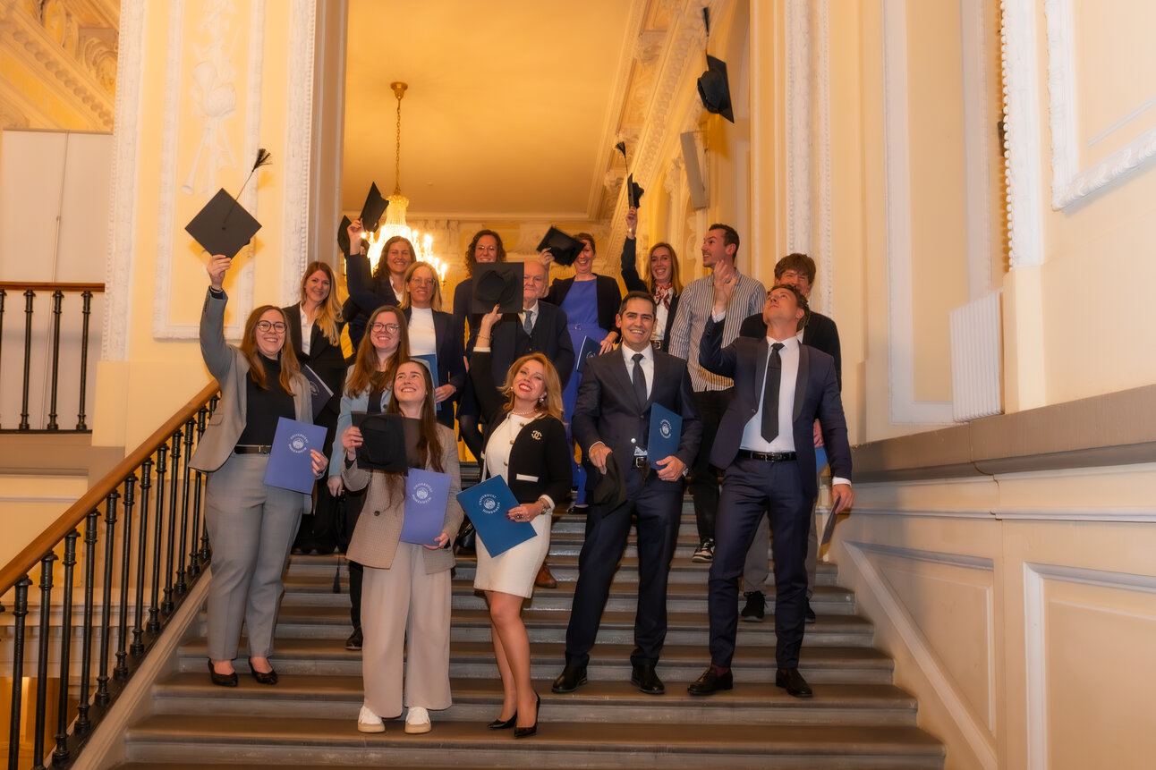 Foto: Eine Gruppe von Personen in formeller Kleidung steht auf einer Treppe in einem Gebäude mit dekorativen Wänden. Einige halten blaue Mappen und werfen Hüte in die Luft.